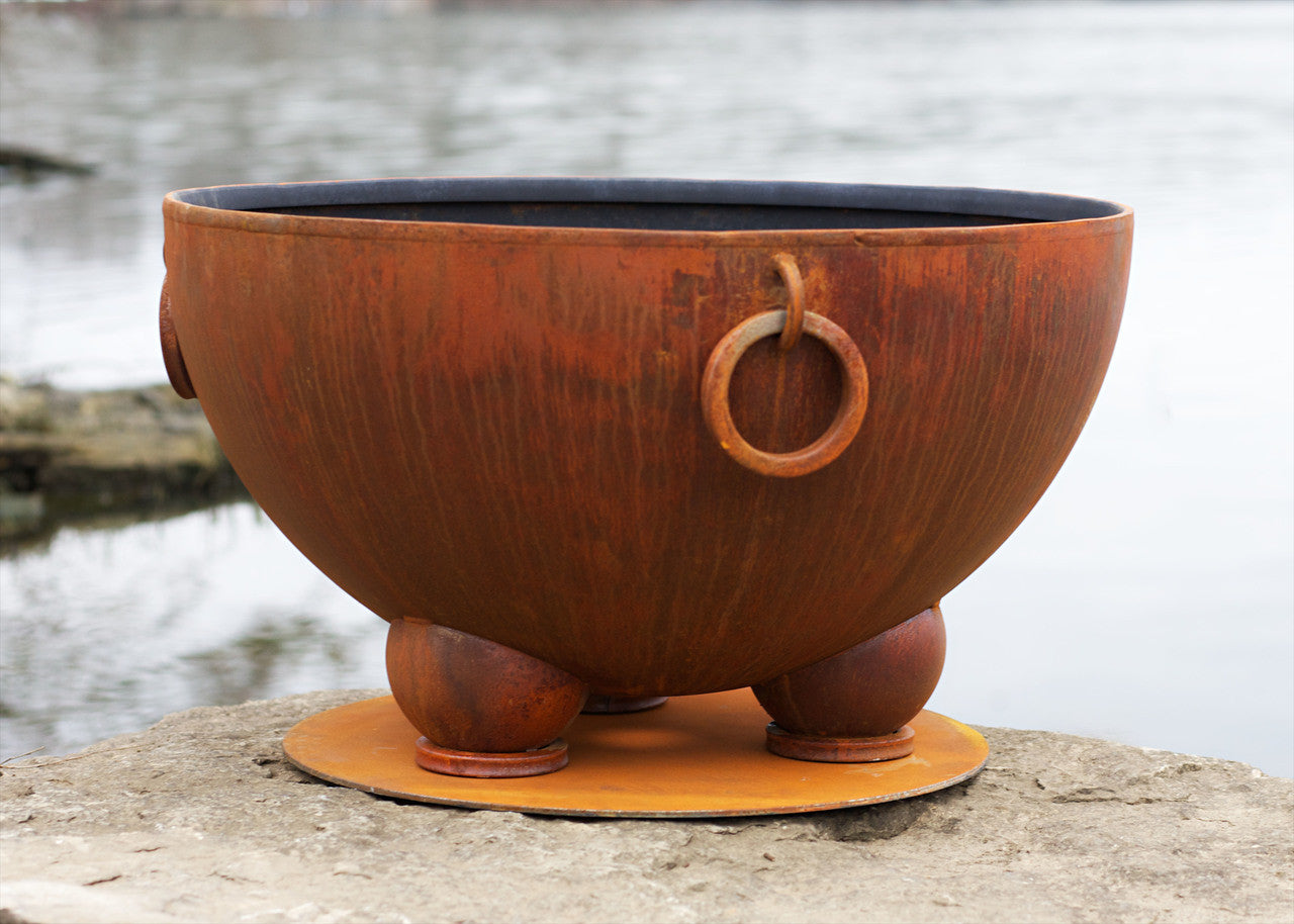 Decorative metal bowl used for wood burning fire pit with a ring handle on a stone surface by a water body