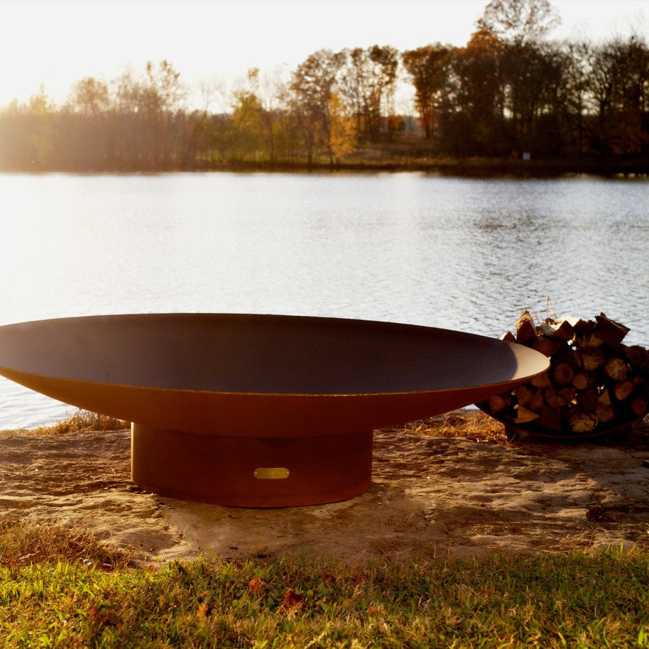 Rust-colored fire pit by a lake with trees in the background