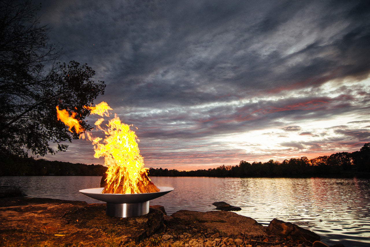 Fire pit by a lake with a dramatic sky during sunset or sunrise.