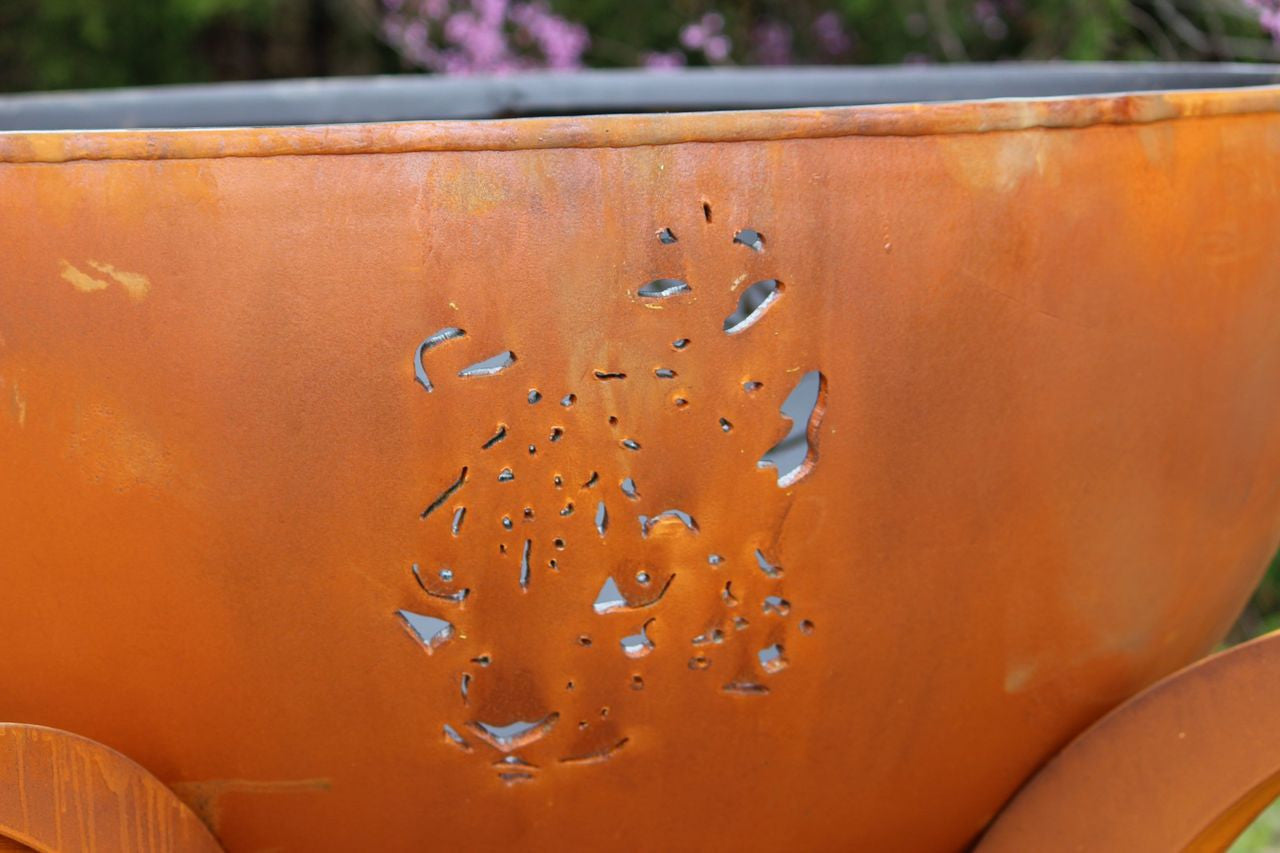 Rust-colored metal fire pit with cut-out design against a blurred natural background