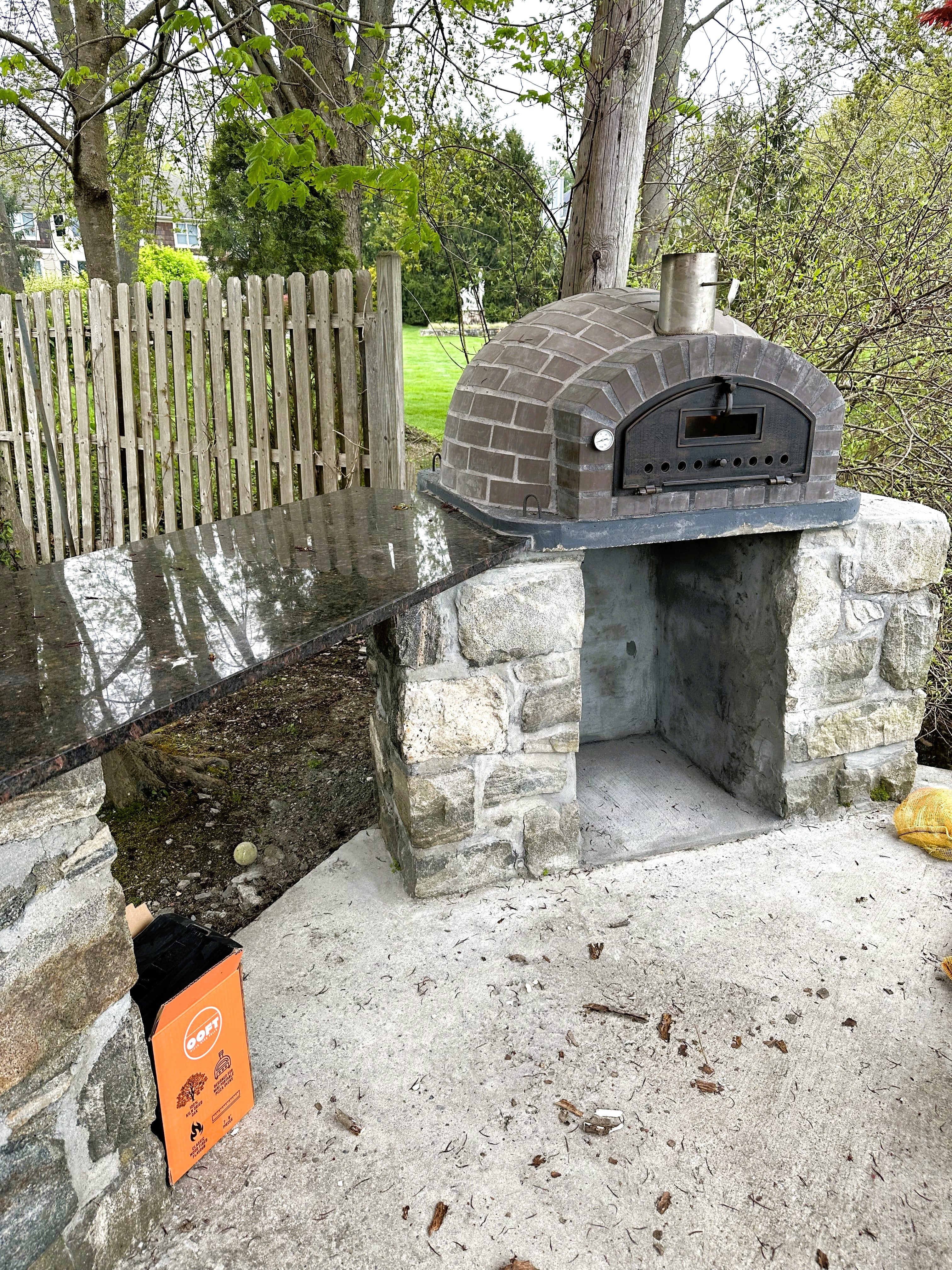 Outdoor stone pizza oven with a stone countertop in a garden setting.