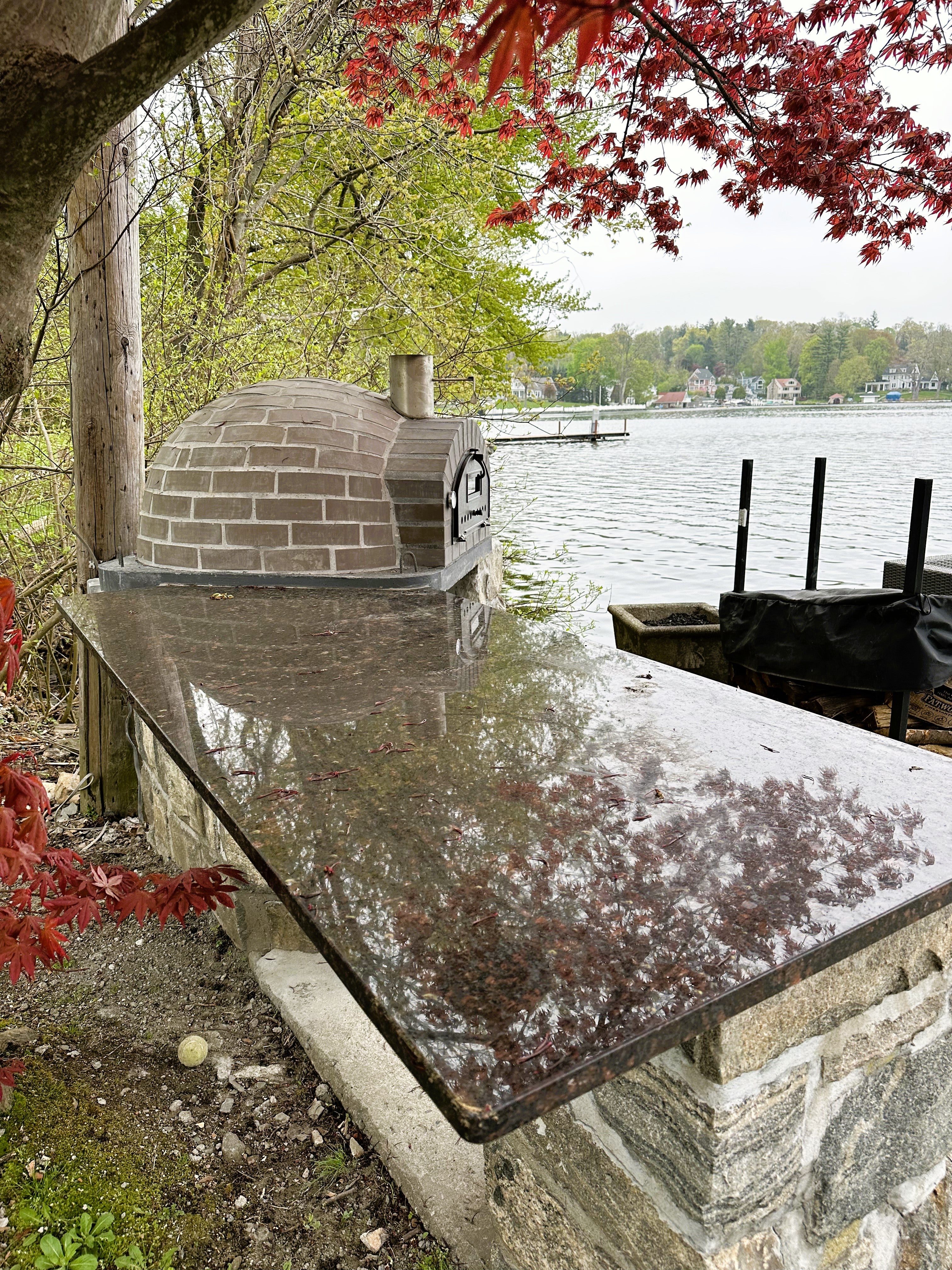 Outdoor stone oven by a lake with trees and water in the background