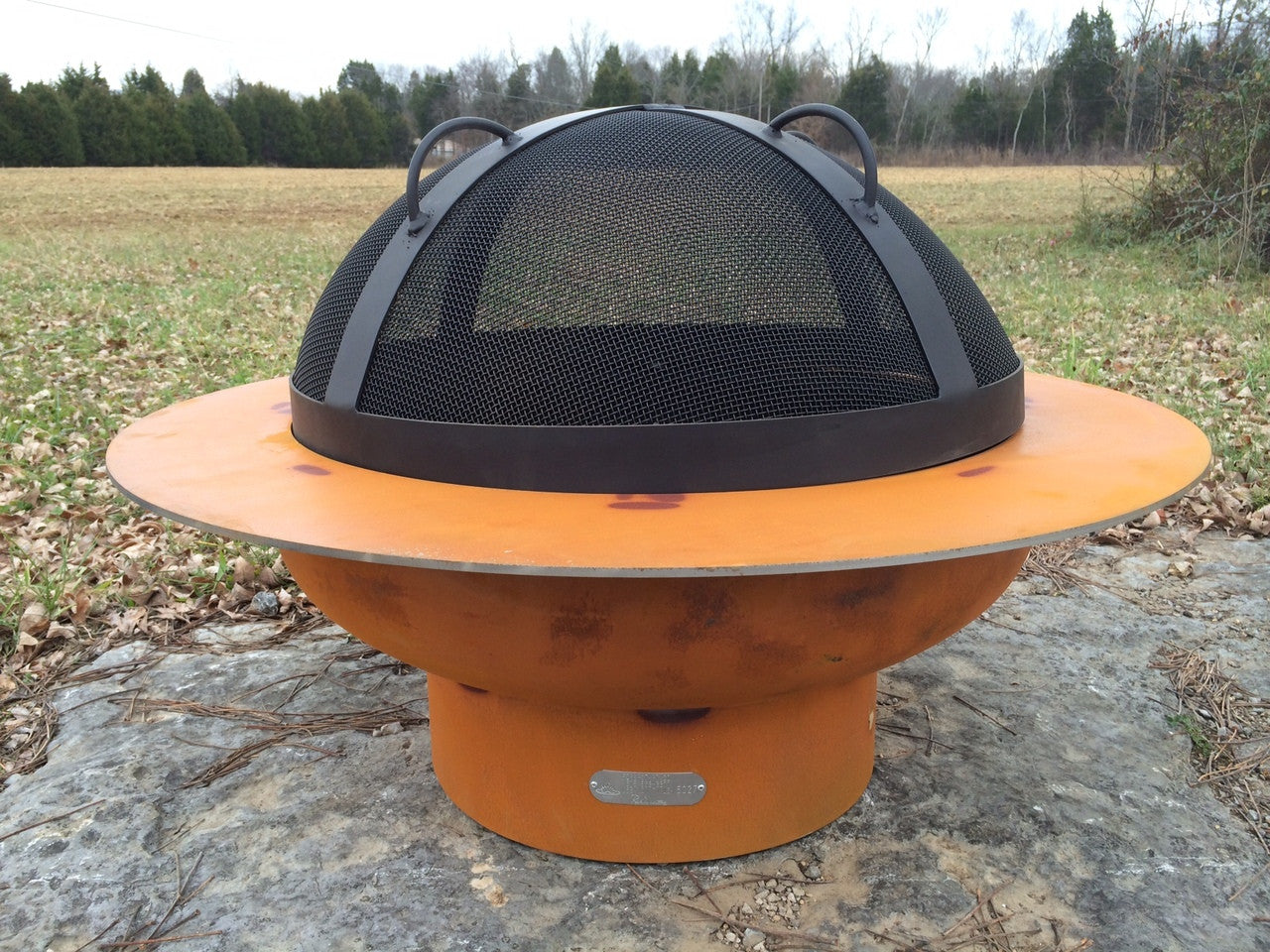Orange metal fire pit with black mesh fire guard cover on a natural outdoor setting