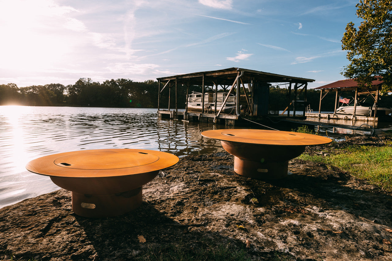 Two fire pits with lids on a lakeside with a dock and trees in the background.