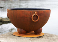 Decorative metal bowl used for wood burning fire pit with a ring handle on a stone surface by a water body