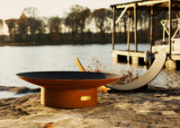 Fire pit outside on a lakeside patio with a dock and trees in the background with firewood unique holder