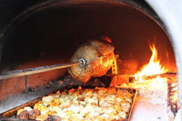 Roast being cooked on a spit inside a brick pizza oven with wood fired flames and glowing embers.