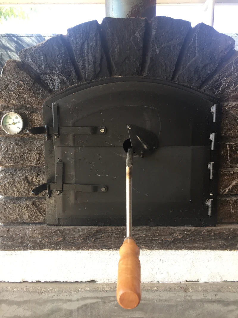 Roast being cooked on a spit inside a brick pizza oven with wood fired flames and glowing embers.